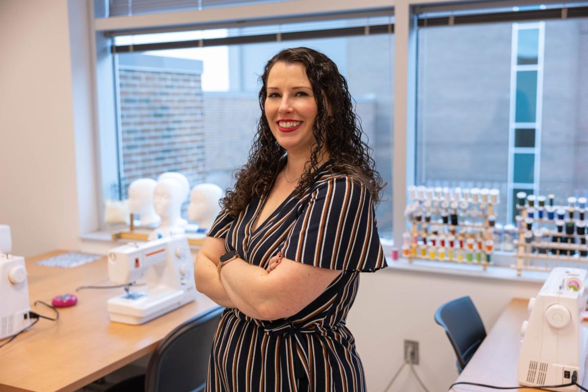 Alt text:  A woman with curly dark hair stands smiling with her arms crossed in a sewing classroom. Behind her are sewing machines on tables, mannequin heads, and racks of colorful thread near large windows.
