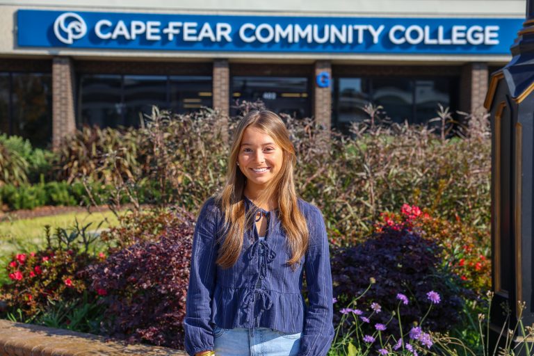 Smiling student standing outdoors in front of the Cape Fear Community College building sign, with landscaped flowers and shrubs in the foreground.
