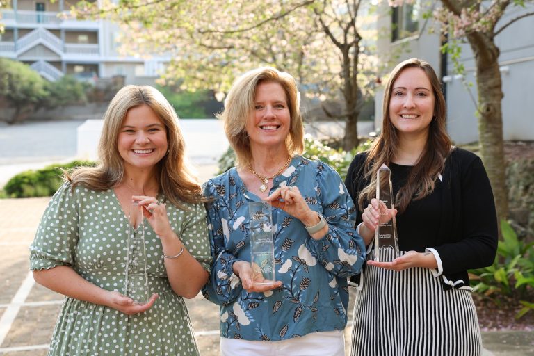 Three women stand outdoors on a sunlit walkway, smiling and holding clear glass award plaques. The woman on the left wears a green polka-dot dress, the center woman wears a blue patterned blouse, and the woman on the right wears a black top with a striped skirt. Trees with blossoms and a residential-style building appear in the background.
