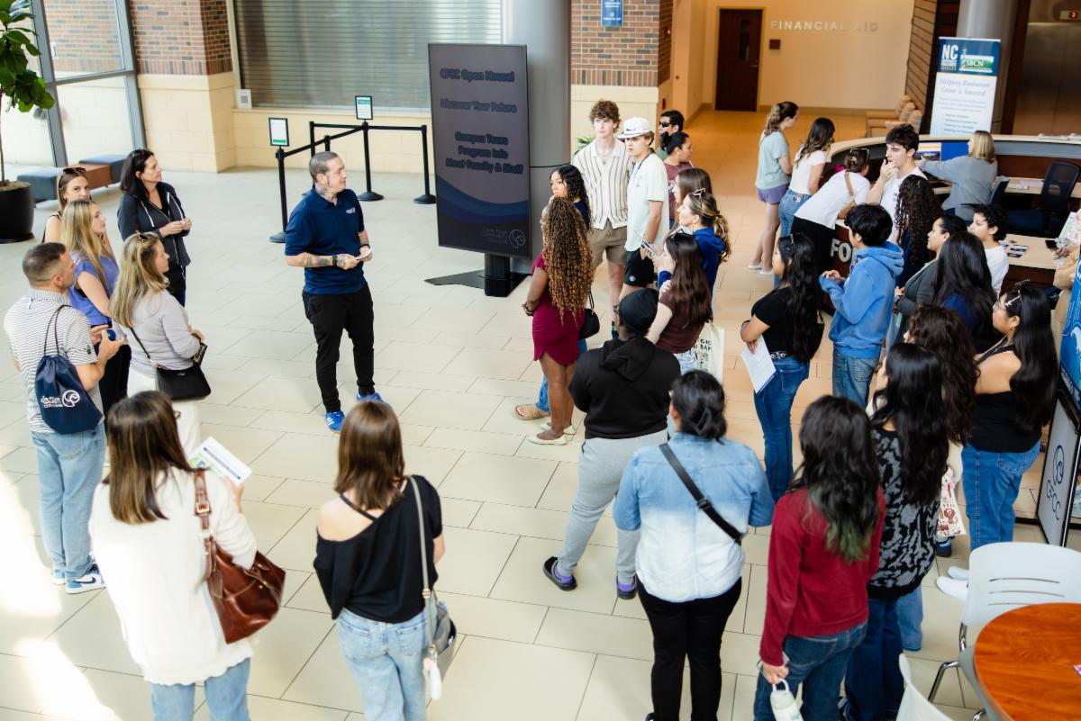 A group of prospective students stands in a lobby listening to a staff member speaking during a Cape Fear Community College open house, with a sign about campus tours and meeting faculty nearby.