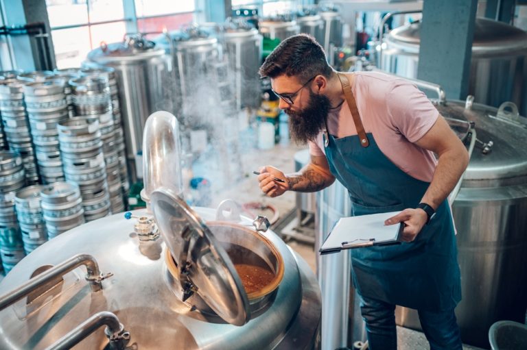 Brewer inspecting a large stainless steel brewing tank inside a brewery, holding a clipboard and checking the contents as steam rises from the open vessel. Rows of metal kegs and additional brewing equipment are visible in the background.