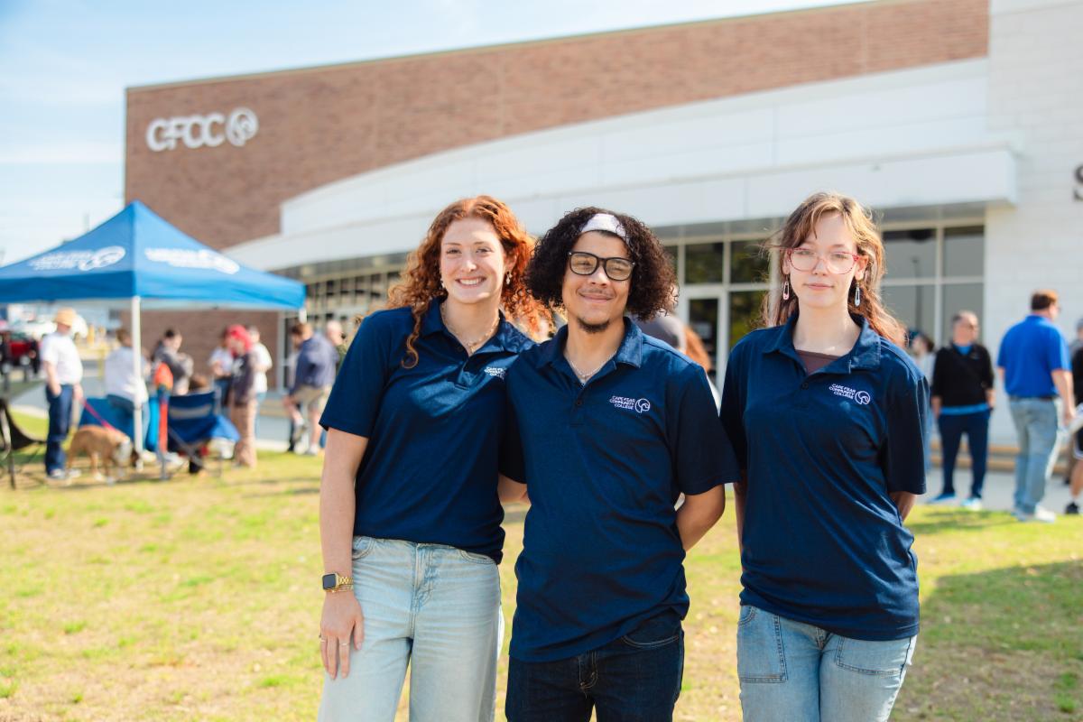Three people wearing navy polo shirts stand together smiling in front of a Cape Fear Community College building during an outdoor event, with a blue tent and small crowd visible in the background.