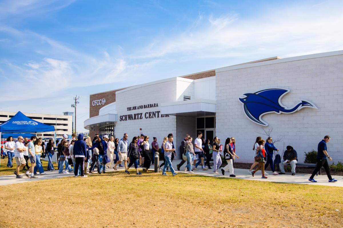 A large group of people walks along a sidewalk toward the Schwartz Center building at Cape Fear Community College during a campus event, with a blue tent set up on the lawn.
