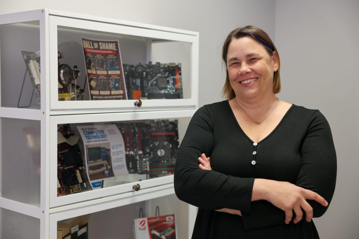 Smiling woman with arms crossed standing beside a display cabinet containing computer hardware and technology-related items in an office or classroom setting.