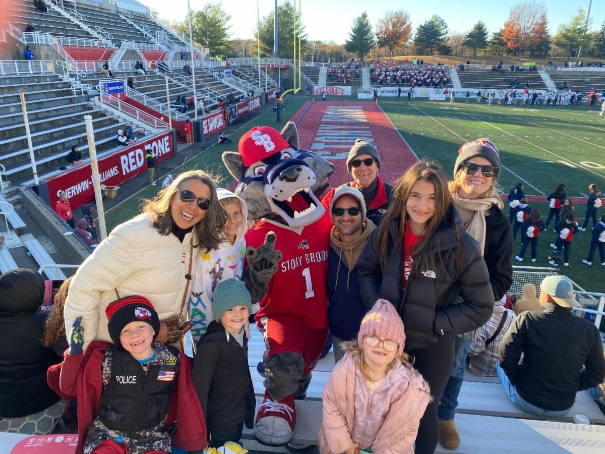 Students with their families on the bleachers with the football field behind them with Wolfie mascot to support the Deans' Challenge football game.