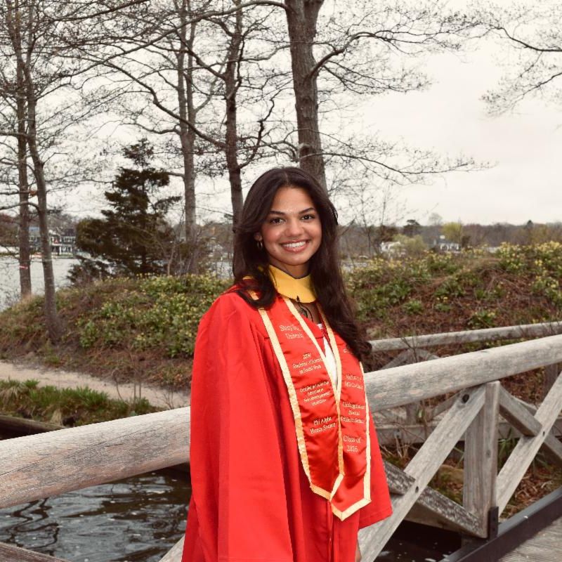 BSW Student in red regalia standing on a bridge overlooking a body of water with trees behind her.