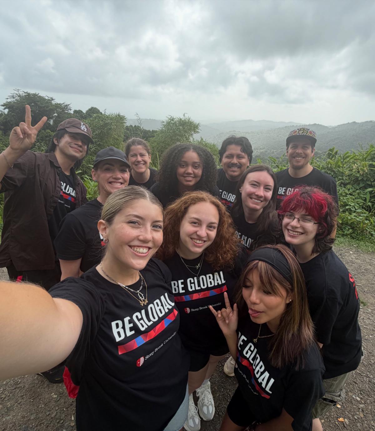Various students wearing black "BE GLOBAL" shirts with smiles and peace signs over a lush tropical landscape.