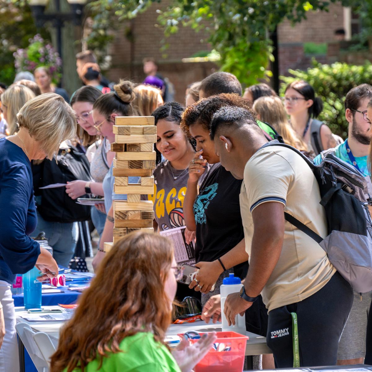 People gather around a table at an outdoor event with a focus on a Jenga like game