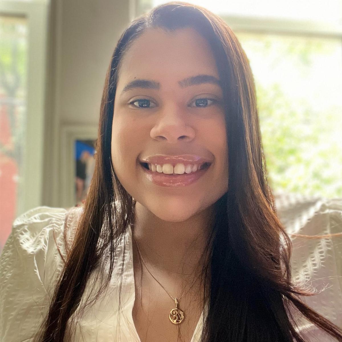 Close-up portrait of a smiling person with long brown hair, wearing a white blouse, dark vest, and pendant necklace.