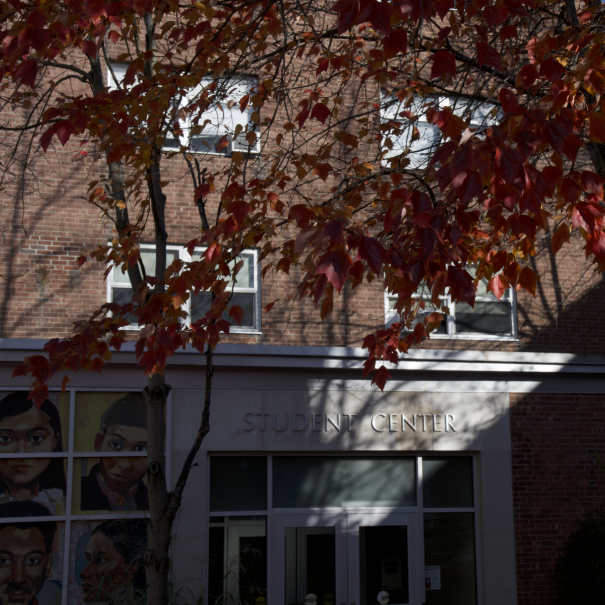 Autumn leaves frame the entrance of a brick student center, with painted portraits in the windows and â€œSTUDENT CENTERâ€ above the doors.