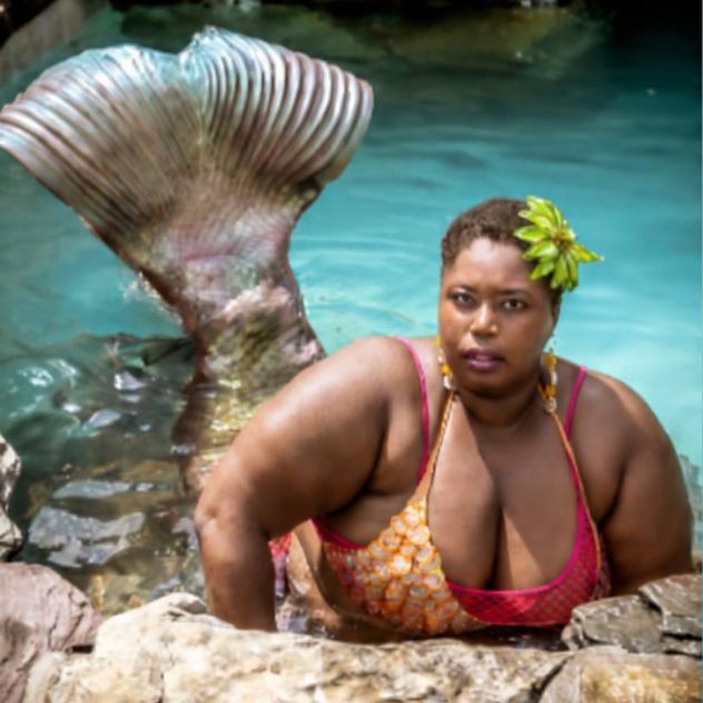 Person in a pink-and-orange mermaid costume poses in turquoise water beside a large ridged tail.
