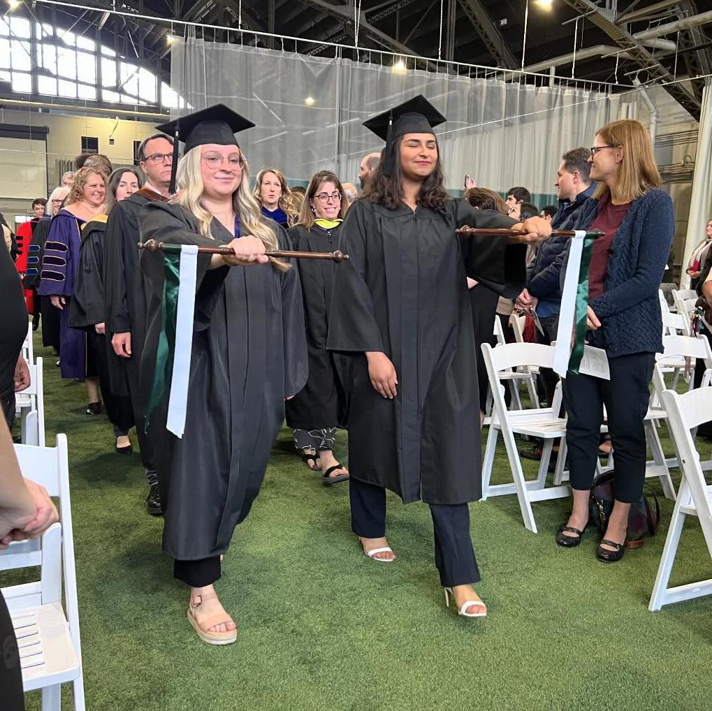 Students in caps and gowns holding a ceremonial staff during an indoor procession