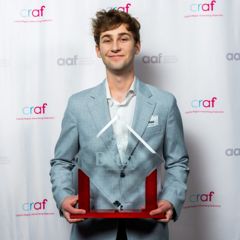 Person in a light gray suit holding an award in front of a branded backdrop.