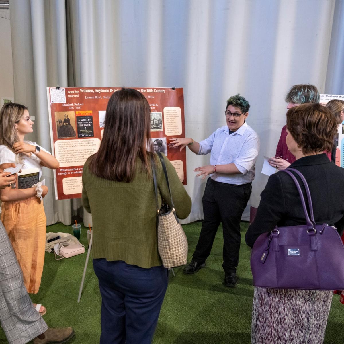 A presenter explains a historical poster to a group of five people in an indoor setting