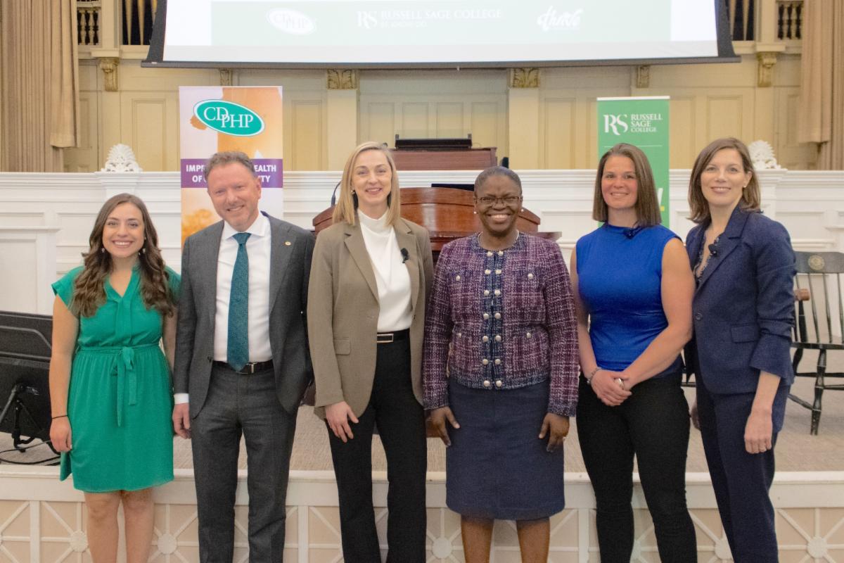 Six people in professional attire standing in line indoors with a podium and banners in the background