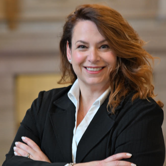 Smiling woman with auburn hair in a black blazer and white shirt, standing with her arms crossed.