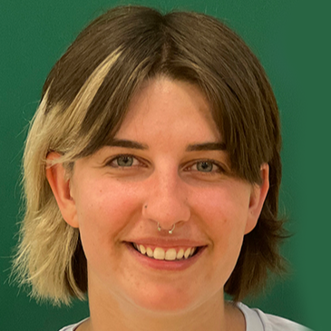 A person with short brown hair and a blonde streak on one side and with a septum piercing smiles in front of a green background