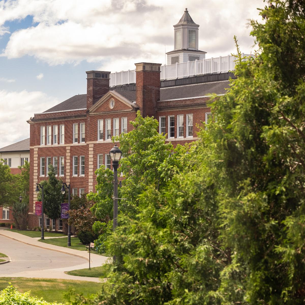 Red-brick campus building with a white cupola, surrounded by trees, lampposts, and purple banners.