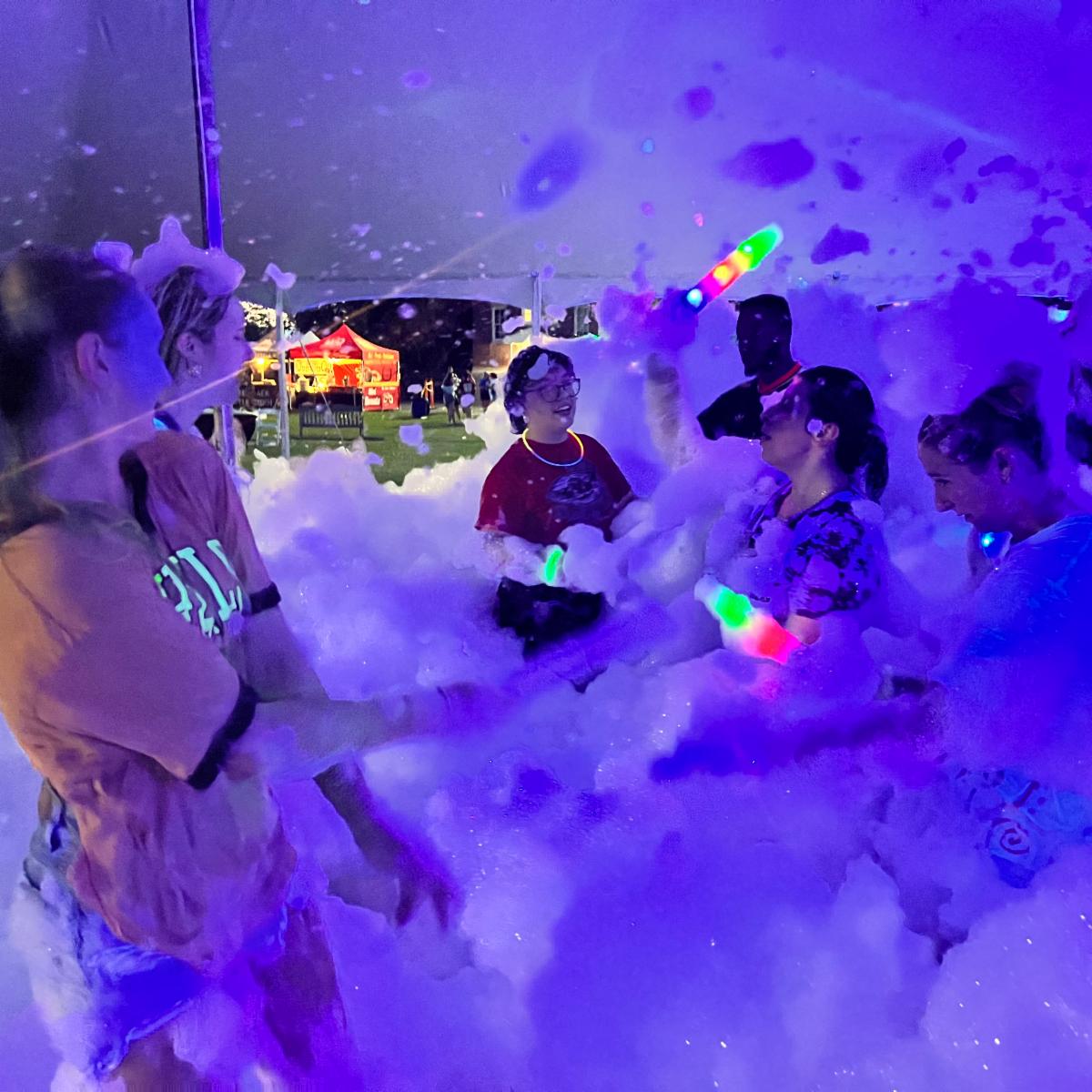People enjoy a blue- and purple-lit foam-filled outdoor event at night.
