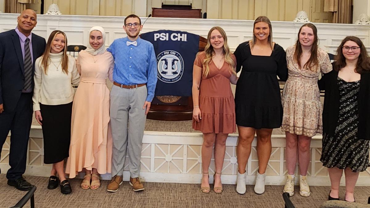 Eight university students pose in front of a podium with a Psi Chi Honor Society banner