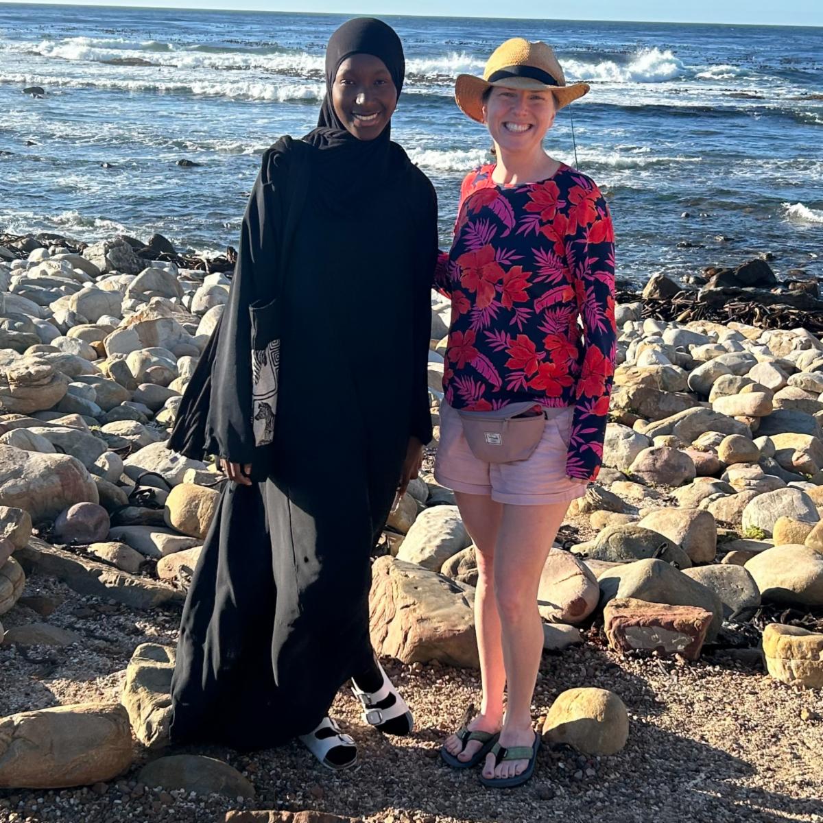 Two women standing together on rocky beach one in traditional black hijab and abaya and and the other in casual colorful clothing with a straw hat with the ocean in the background.