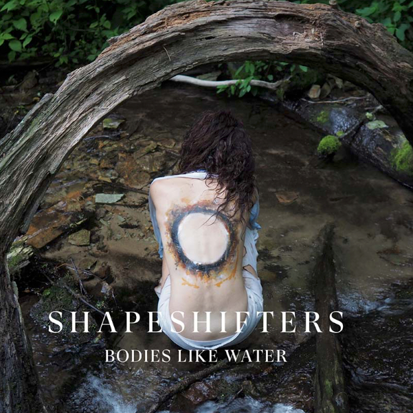 Person seated in a shallow forest stream beneath an arched tree trunk, with a painted circular design on their back. Text reads, â€œSHAPESHIFTERS: BODIES LIKE WATER.â€