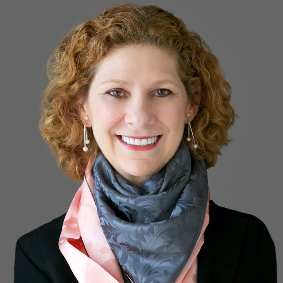 Head-and-shoulders portrait of a smiling woman with curly auburn hair, pearl earrings, a dark blazer, and layered blue-gray and pink scarves against a gray background.