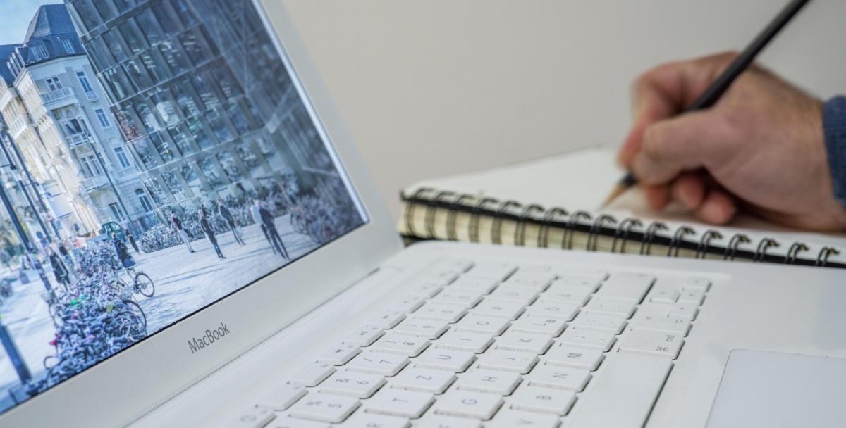 a hand holding a pencil while writing in a notebook set beside an open laptop