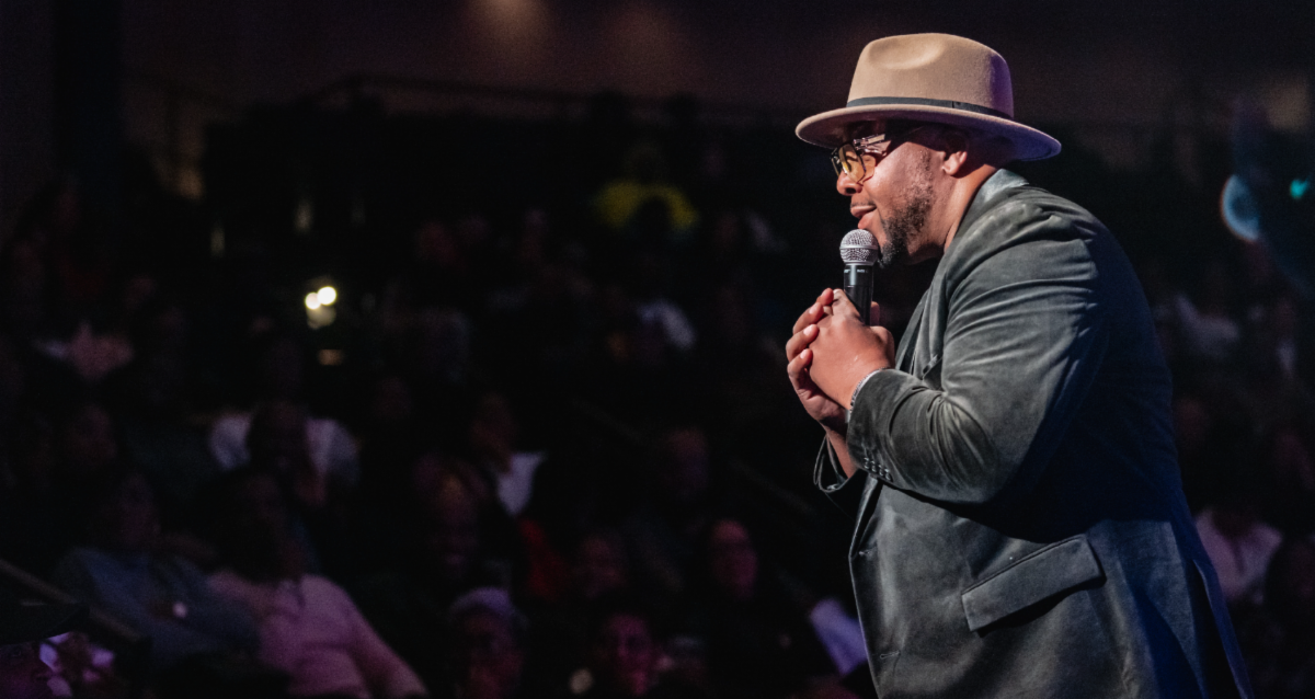 Performer speaks into a microphone on stage in front of a seated audience.