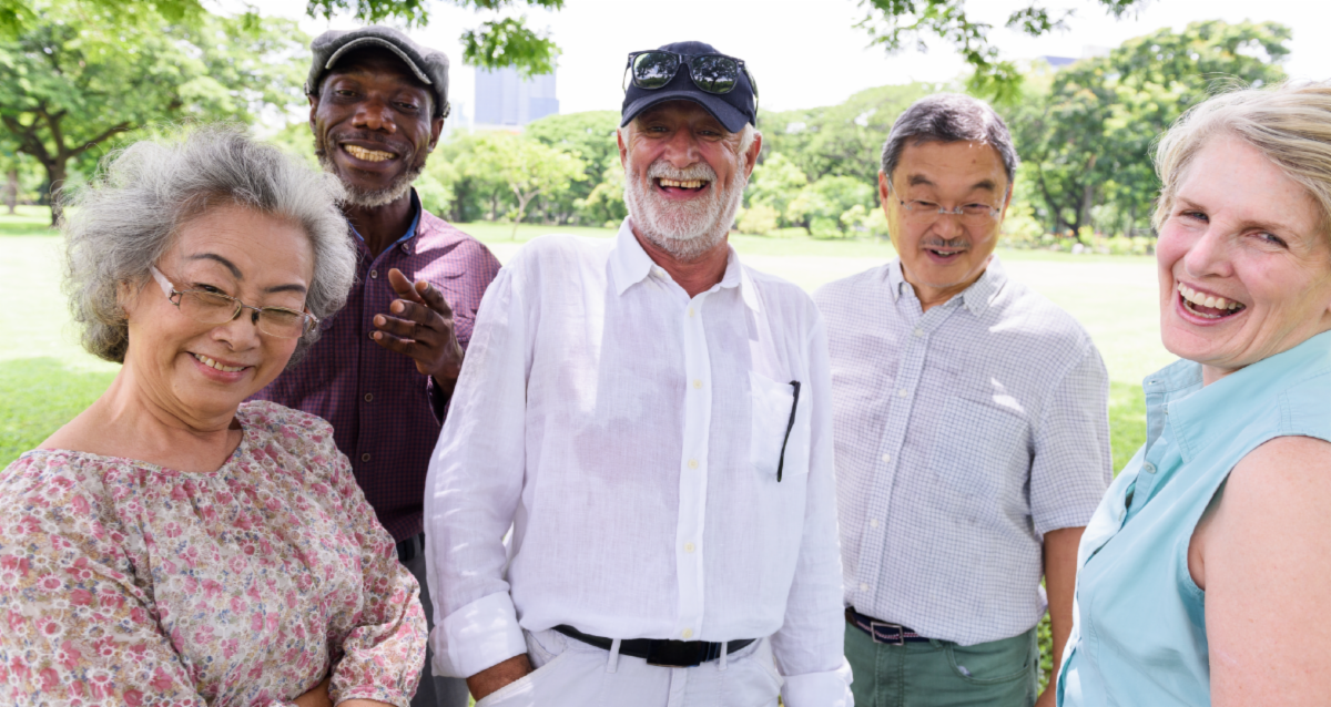 Photo of five smiling older adults from diverse backgrounds standing together outdoors in a sunny park setting.