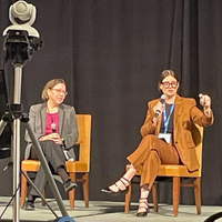 Two people sitting on stage. One of them talking in a microphone