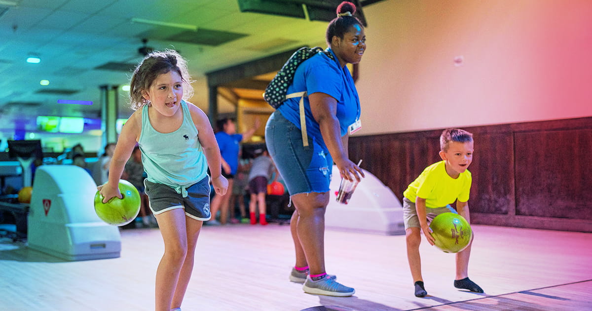 Two campers and a counselor look down a bowling alley lane while the campers prepare to throw their balls.