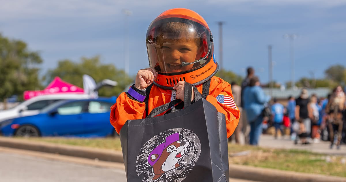 Child at a Halloween event dressed in an orange spacesuit and helmet, and carrying a purple bag.