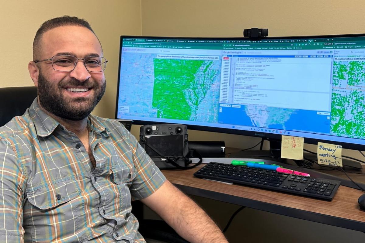 A man smiles with a large computer screen showing a map of Arkansas in the background