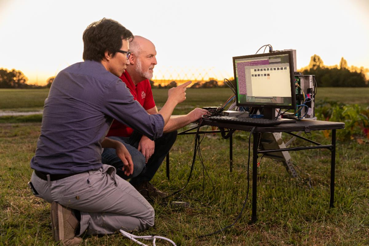 Two men crouch looking at a computer screen in an agricultural field.
