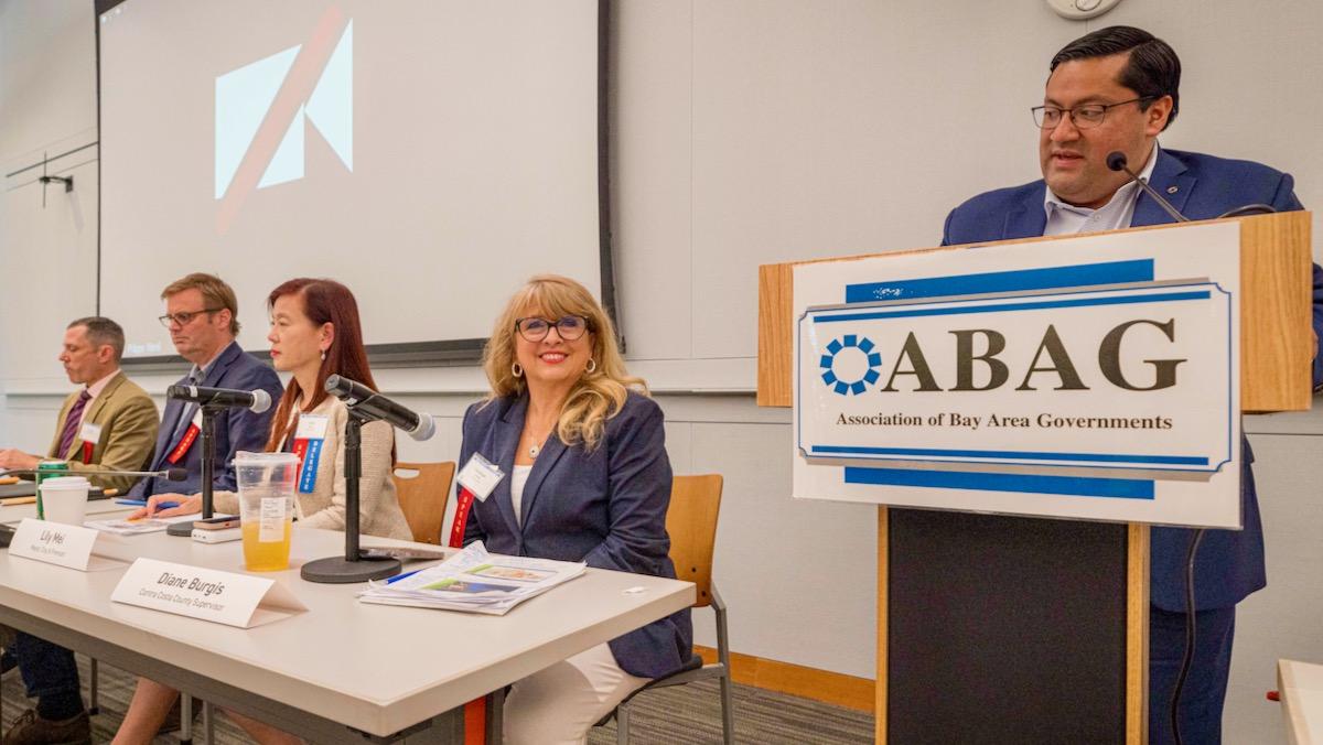 Supervisor Burgis sits at a microphone while Berkeley Mayor Jesse Arreguin speaks at a podium