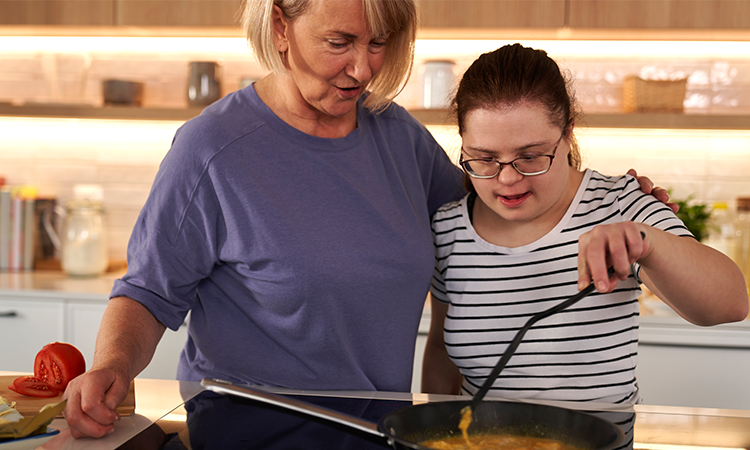Two women cook together in a kitchen, with one stirring food in a frying pan while the other stands beside her with an arm around her shoulder.
