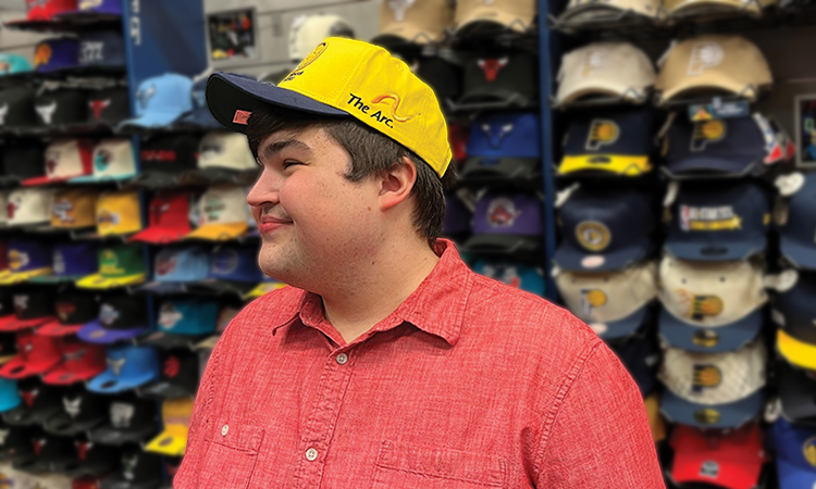 Young man in a red shirt wearing a yellow The Arc hat standing in a store with shelves filled with sports caps in various colors and team logos
