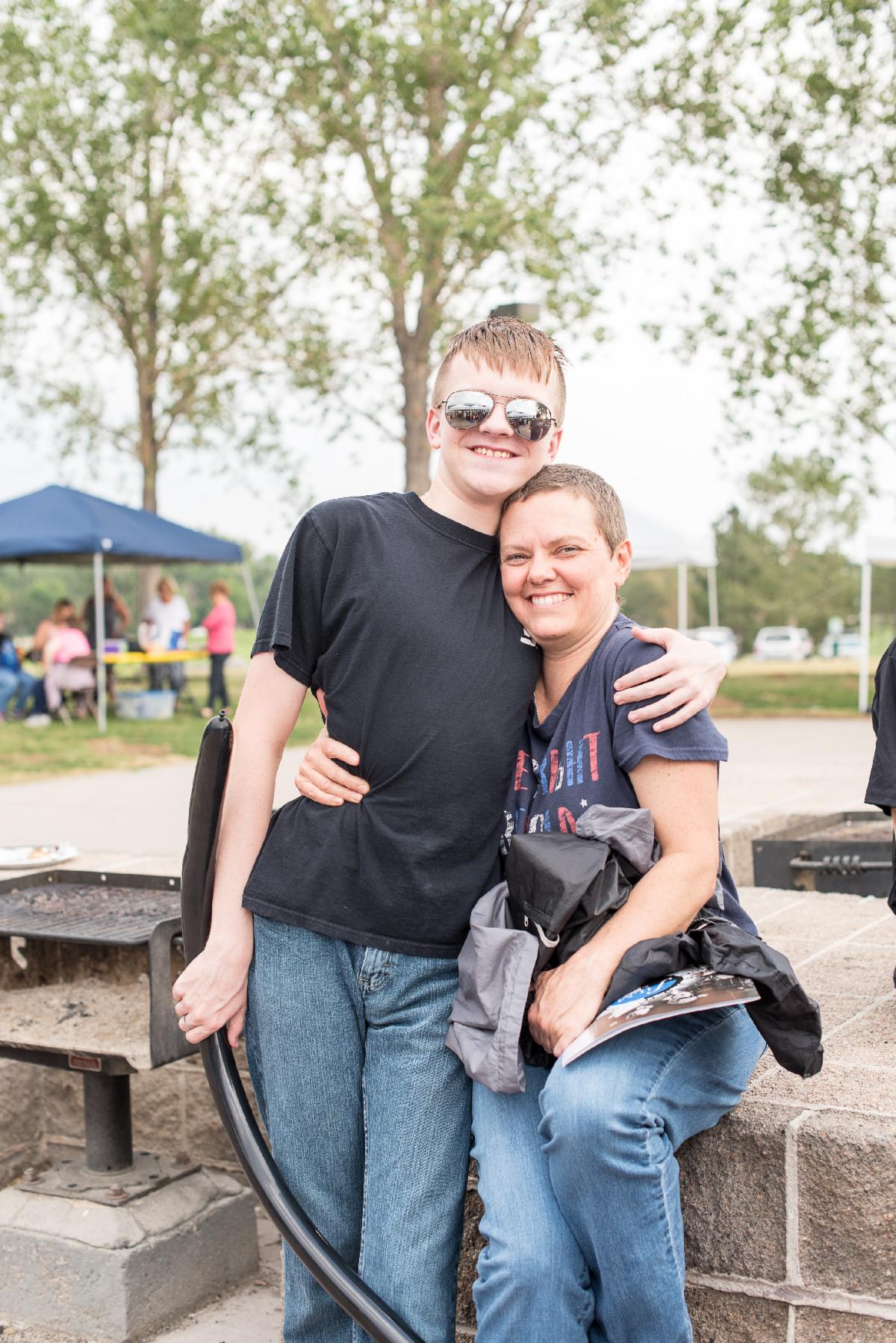 Two people smile and pose together outdoors, with one wearing sunglasses and an arm around the other, who is seated on a low wall.
