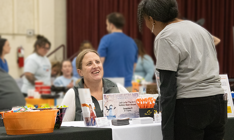 A staff member sits behind a table speaking with a visitor at a community resource fair. Informational materials and giveaway items are displayed on the table while others gather in the background.