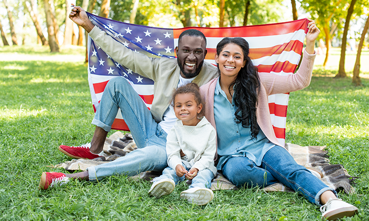 A family sits on a blanket in a park, smiling and holding up an American flag behind them.