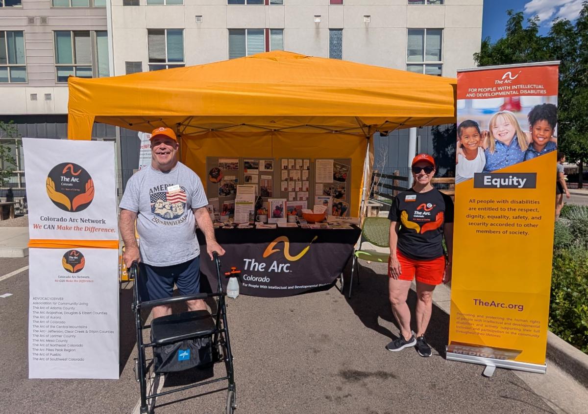 Two people standing beside The Arc Colorado booth under an orange tent