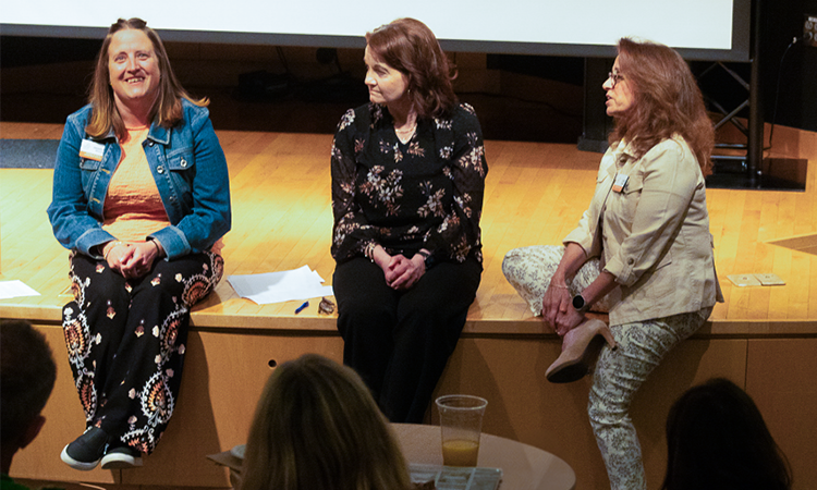 Three people seated on a stage during a panel discussion, facing an audience; papers and a cup are visible nearby.