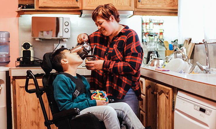 An adult pours a drink for a child using a wheelchair in a kitchen. The child holds a colorful sensory toy.