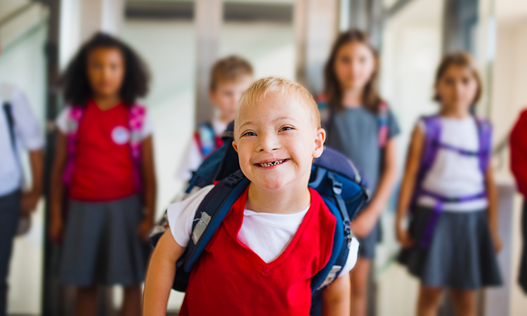 Smiling boy with Down syndrome wearing a red shirt and backpack stands in front of classmates