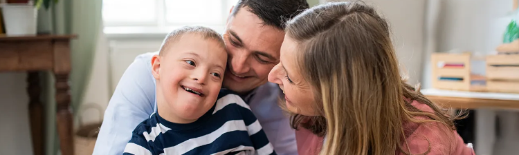 A young boy in a striped shirt smiles while embraced by two adults who lean close with affection