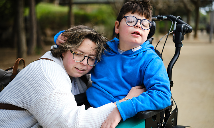Adult supports a child wearing glasses who sits in a wheelchair outdoors on a park path.