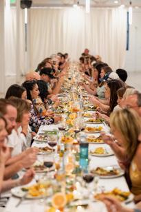 A long banquet table is filled with people enjoying dinner together in a warmly lit setting