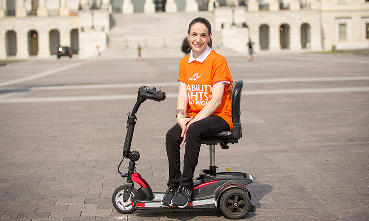 A person wearing an orange The Arc T shirt sits on a mobility scooter smiling in front of a large white building with columns