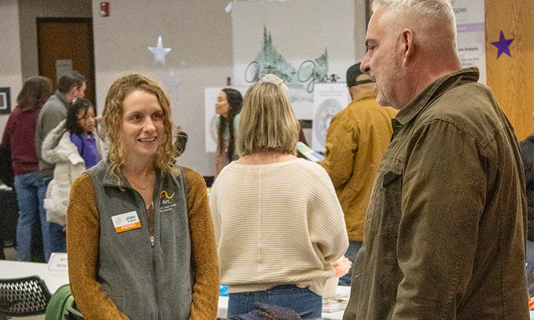 Two adults talk at a community event indoors, with tables and other attendees in the background.